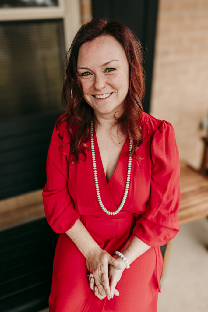 Kirkwood School Board candidate Laura Hepburn sits on her front porch, hands clasped in her lap. She is smiling toward the camera and wearing a Pioneer red dress with a beaded necklace and bracelet.